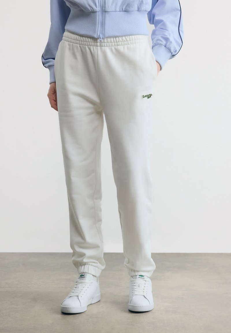 Άτομο που φοράει λευκή φόρμα Lacoste και λευκά αθλητικά παπούτσια Lacoste, στέκεται σε γκρίζο πάτωμα με λευκό φόντο.