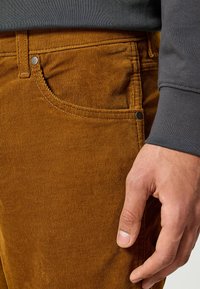 Bruine corduroy broek met een geribbelde textuur, voorzien van een voorzak, een metalen knoop en zichtbare stiksels. Grijze mouw gedeeltelijk zichtbaar.
