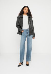 Chaqueta de cuero negra, top gris, pantalones vaqueros de pierna ancha azul claro y tacones altos negros. La modelo está de pie frente a un fondo blanco liso.
