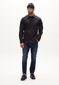 Cămașă neagră cu nasturi, cu un model geometric, asortată cu blugi albaștri și pantofi negri. Modelul poartă o căciulă din lână închisa la culoare.