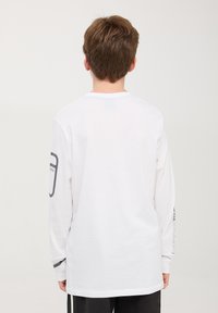 T-shirt bianco a maniche lunghe in cotone. Presenta un collo rotondo, una linea dritta e un dettaglio testuale grafico sulla manica sinistra.