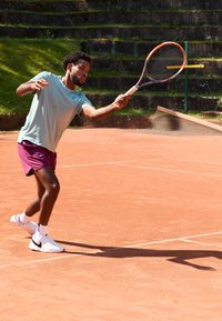 Tennis-Spieler in hellblauer Shirt und kastanienbraunen Shorts, der einen Ball mit einem Schläger auf einem Sandplatz schlägt und weiße Turnschuhe trägt.
