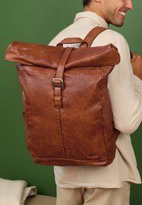 Pruunist nahast seljakott, millel on rullkattega disain, reguleeritav rihm ja küljetasku. Sile tekstuur loomuliku viimistlusega.