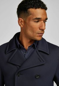 Marineblauer doppelt knöpfbarer Mantel mit großem Schalkragen, strukturiertem Stoff und schwarzen Knöpfen, kombiniert mit einem dunkelblauen Hemd darunter.