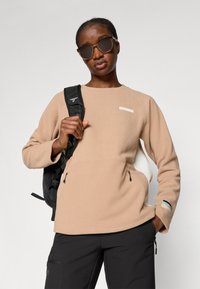 Beige fleece neule, jossa on teksturoitu kangas, kaksi vetoketjullista taskua ja brändilogo-patch. Musta reppu, jossa on säädettävät hihnat.