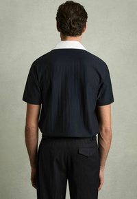 Cămașă scurtă cu mânecă, de culoare bleumarin și cu guler alb, asociată cu pantaloni în dungi închise, privită din spate. Textură netedă a țesăturii.