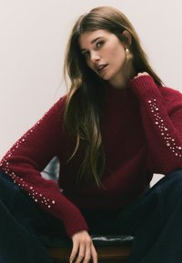 Röd stickad tröja med hög krage, med detaljer på ärmarna. Modellen har på sig mörka jeans och sitter mot en mjuk yta.