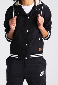Sort vindjakke med en hvid foret hætte, trykknapper og stribede manchetter, samt mørke sweatpants med et Nike-logo.