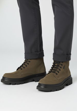 Bruine suède enkellaarsjes met zwarte rubberen zolen en een veterontwerp. Heeft een afgeronde neus en een gestructureerd bovenwerk, gecombineerd met donkere broeken.