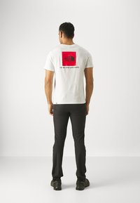 Witte t-shirt met een rode vierkante grafiek waarop het "The North Face"-logo en de tekst "NEVER STOP EXPLORING" op de achterkant staan. Zwarte broek en schoenen.