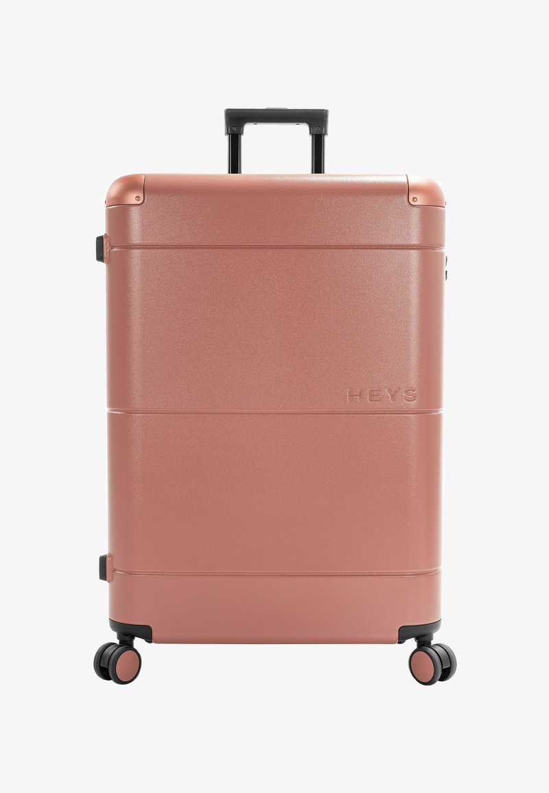Valise rigide couleur or rose avec quatre roues pivotantes et une poignée télescopique noire, nom de la marque "HEYS" embossé à l'avant.