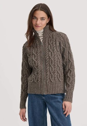 Bruin gebreid sweater met hoge kraag, voorzien van een kabelpatroon, rits sluiting en ribbel zoom, gedragen over een lichte coltrui.