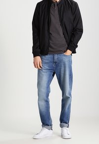 Svart bombarjacka över en grå polo, blå avslappnade jeans och vita sneakers. Materialet verkar slätt med en avslappnad design.