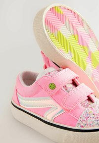 Zapatillas de ante rosa con punta brillante, detalles blancos y un botón sonriente. Cuentan con cintas de velcro y suela de goma texturizada con un patrón en zigzag.