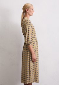Lang beige jurkje met blauwe geometrische patronen, heeft een aansluitende taille en pofmouwen, gemaakt van een gladde stof, met een midi-lengte.