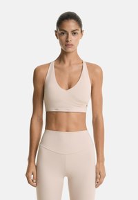Light beige sports bra with a deep V-neck, wide straps, and a crossover front design. Smooth fabric and no visible hardware.