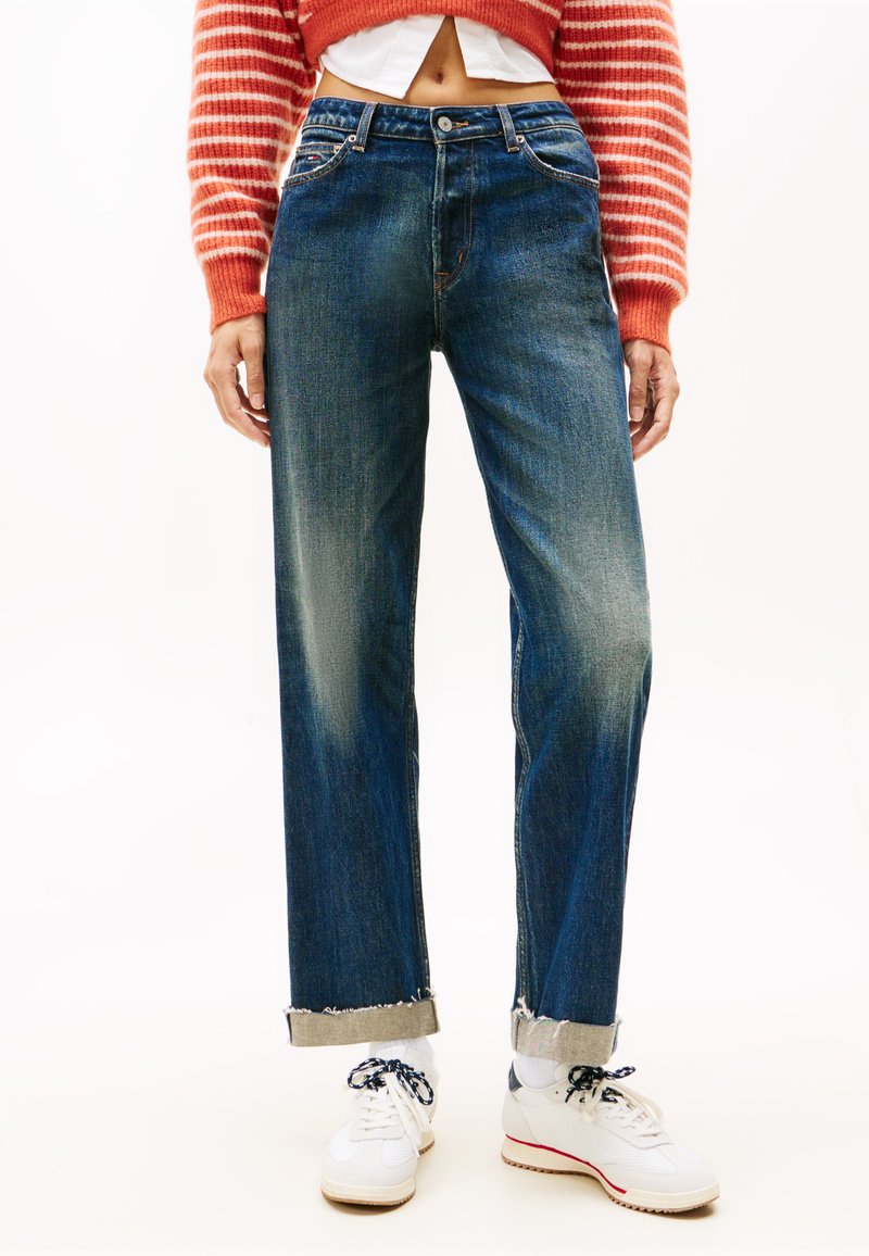 Kék farmernadrág egyenes lábbal, nyers szegéllyel és öt zsebbel. Piros csíkos cropped pulóverrel és fehér sportcipővel viselve.