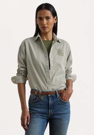 Camicia a righe verde e bianca, con un logo sul petto, abbinata a uno strato inferiore verde, jeans blu e una cintura marrone.