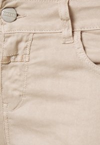 Pantalons beige avec une texture lisse, dotés d'une poche avant et d'un détail en bouton argenté avec l'inscription "CLOSED". Couture adjacente visible.