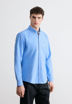 Jeune homme portant une chemise bleu clair à boutons avec manches longues et un pantalon noir, debout devant un fond clair uni.