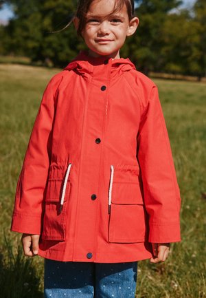 Dievča v červenej bunde s kapucňou s bielymi sťahovacími šnúrkami a modrých nohaviciach stojí vonku na tráve s stromami v pozadí.