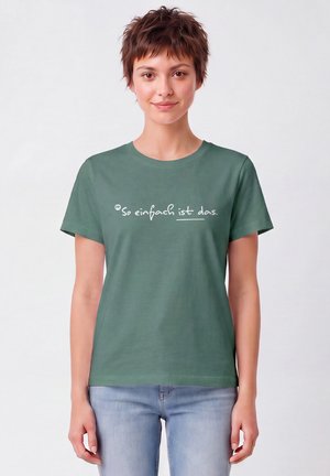 Jong persoon met kort haar die een groen T-shirt draagt met de Duitse tekst "So einfach ist das" en lichtblauwe spijkerbroek tegen een effen achtergrond.