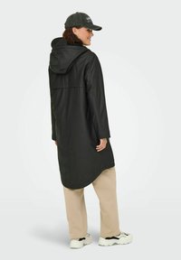 Frau in einem schwarzen Kapuzen-Regenmantel, beige Hosen, weißen Sneakers und einer dunklen Mütze, die sich leicht zur Seite dreht, vor einem einfarbigen Hintergrund.