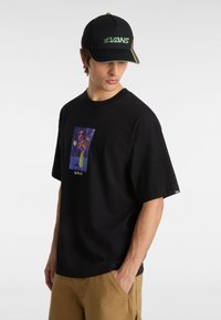 Schwarzes Baumwoll-T-Shirt mit kurzen Ärmeln, das ein buntes Blumenmotiv und das "VANS"-Logo am unteren Rand zeigt. Getragen mit beigen Cargo-Shorts.