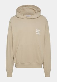 Beige mikina s kapucí vyrobená z měkké tkaniny. Obsahuje přední kapsu a vytisknuté logo "Calvin Klein Jeans" v bílé barvě na levé straně.