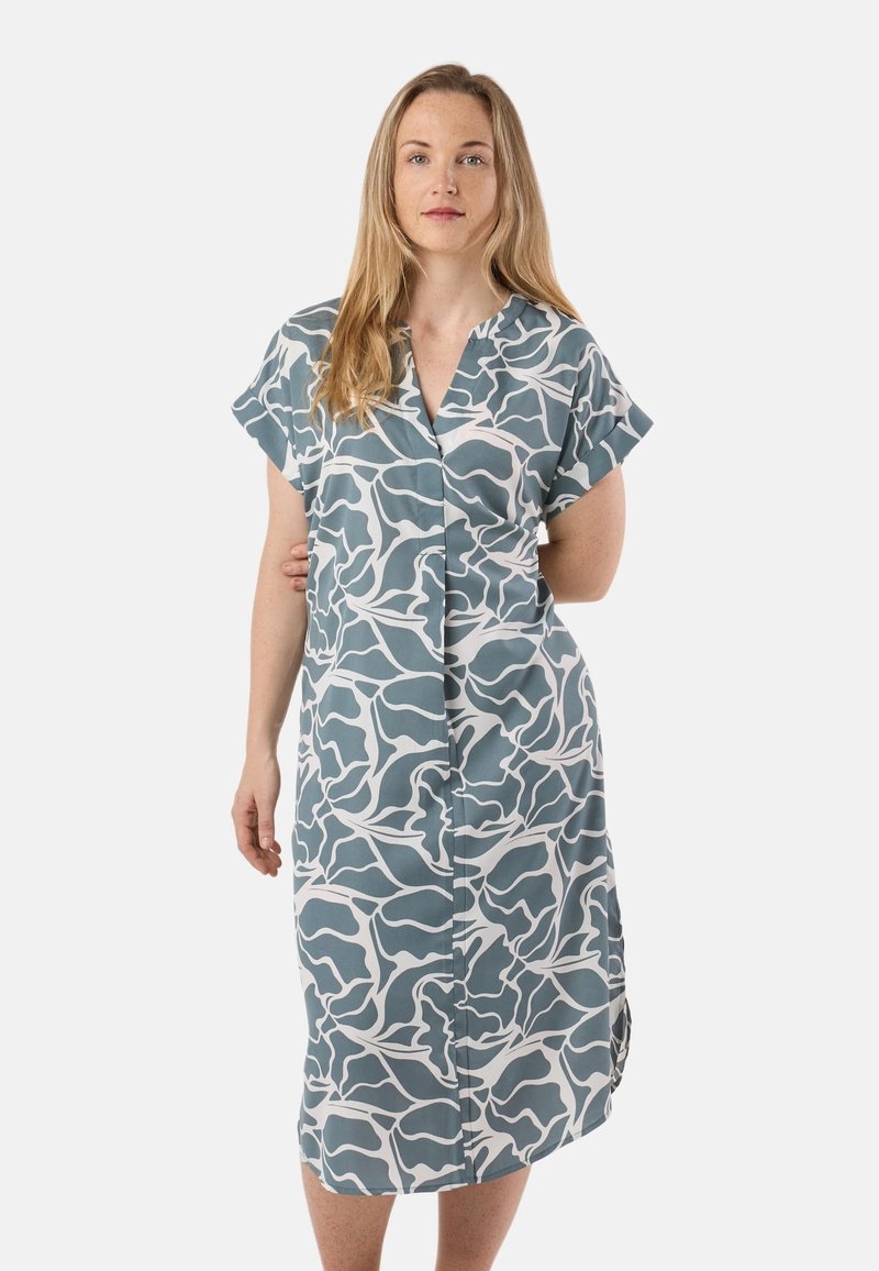 Femme aux longs cheveux blonds portant une robe mi-longue bleu et blanc à motifs abstraits avec des manches courtes retroussées, debout sur un fond blanc.
