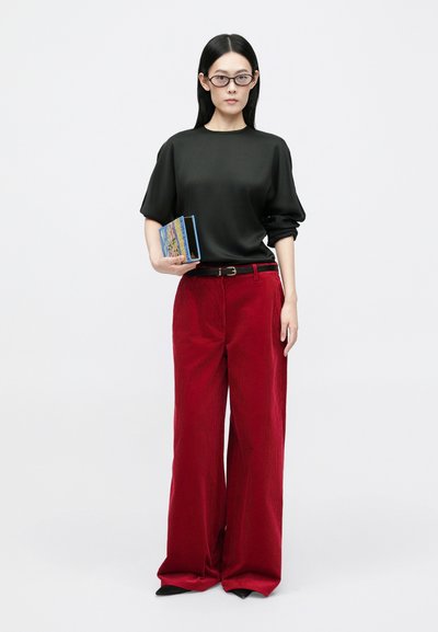 Μαύρη κοντομάνικη μπλούζα, φαρδιά κόκκινα τζιν παντελόνια, μαύρη ζώνη, κρατώντας ένα μπλε βιβλίο με πολύχρωμο εξώφυλλο, απλό λευκό φόντο.