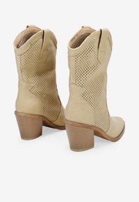 Botas de tobillo de ante perforado beige con tacones apilados y tiradores en la parte superior, que presentan una puntera lisa y paneles laterales con patrón.