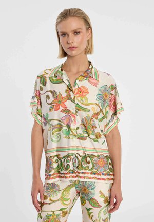 Frau trägt ein locker sitzendes, kurzärmeliges Hemd mit floralem Muster und passende Hosen in Pastellfarben vor einem einfarbigen Hintergrund.