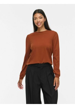 Femme aux longs cheveux foncés portant un haut manches longues couleur rouille et un pantalon large noir plissé, souriante sur un fond uni.