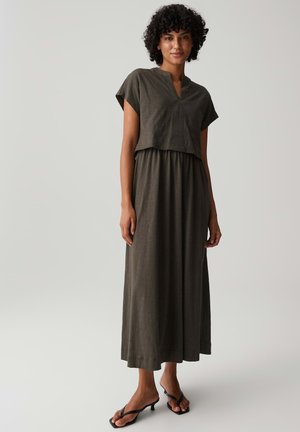 Frau mit lockigen Haaren, die ein dunkelolivfarbenes kurzärmeliges Oberteil und einen passenden langen Rock trägt, steht barfuß auf einem einfarbigen hellen Hintergrund.