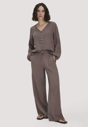 Frau trägt ein lockeres, knöpfbares, langärmeliges Top in Taupe und weite Hose mit Kordelzug, dazu schwarze Flip-Flops, steht mit den Händen in den Taschen.