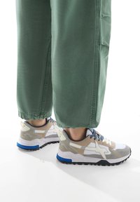 Löparskor med en vit, grå och beige ovandel, blå accenter och en strukturerad sula, kombinerade med lösa gröna byxor.