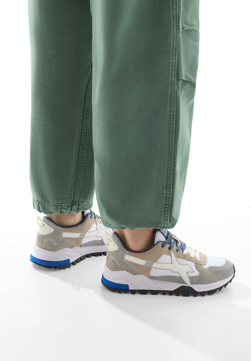 Löparskor med en vit, grå och beige ovandel, blå accenter och en strukturerad sula, kombinerade med lösa gröna byxor.