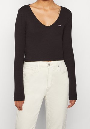 Person wearing a black long-sleeve V-neck cropped top and high-waisted off-white pants against a plain background.