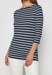 Mujer vistiendo una camisa de mangas largas a rayas horizontalmente en azul marino y blanco, y pantalones blancos con bordado floral, de pie contra un fondo gris.