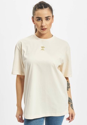 Camiseta oversized color crema hecha de tejido suave, con un pequeño logo dorado en el pecho y mangas cortas. Combina con jeans azules.