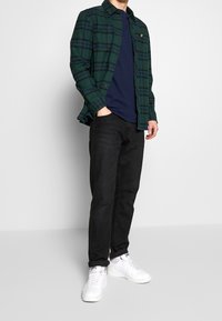 Chemise à carreaux verte et bleue superposée sur un t-shirt bleu marine, assortie à un jeans noir et des baskets blanches, avec un design à boutons.