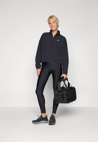 Chaqueta negra corta, leggings negros y zapatillas deportivas negras. El modelo sostiene una bolsa de gimnasio negra, con cierre de cremallera y acentos texturizados.