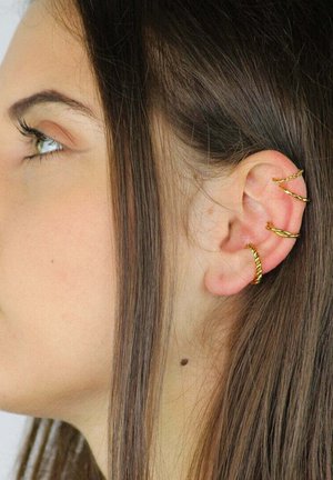 Złote metalowe kolczyki na uchu z zakręconymi wzorami i różnymi rozmiarami kółek, na tle prostych brązowych włosów.