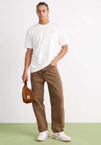 Jovem com cabelo trançado, vestindo uma t-shirt branca da New Balance, jeans castanhos, tênis brancos e laranja, segurando uma bolsa castanha da Carhartt.