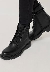 Čierne kožené kotníkové topánky s textúrovaným povrchom, šnurovaním vpredu, bočnými zipsami a tvrdými podrážkami. Navrhnuté pre trvácnosť a trakciu.
