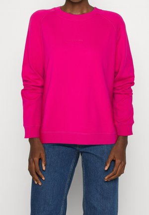Person wearing a bright pink long-sleeve sweatshirt and blue jeans, standing against a plain white background.