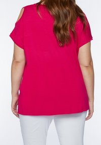 Woman with long brown hair wearing a bright pink cold-shoulder top and white pants, shown from the back against a plain background.