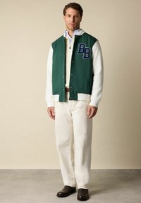 Man in een groen en wit varsityjack met "BB"-patch, crèmekleurige trui, witte broek, bruine riem en donkerbruine schoenen, staande tegen een beige muur.