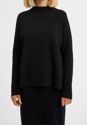 Personne aux cheveux blonds portant un haut à manches longues noir uni et une jupe noire assortie sur un fond blanc.